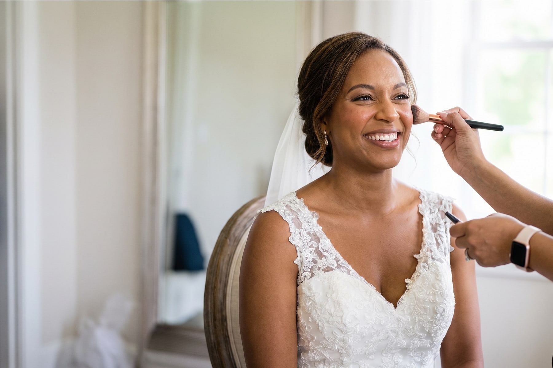 Smiling bride in a lace gown having bridal makeup applied with a brush